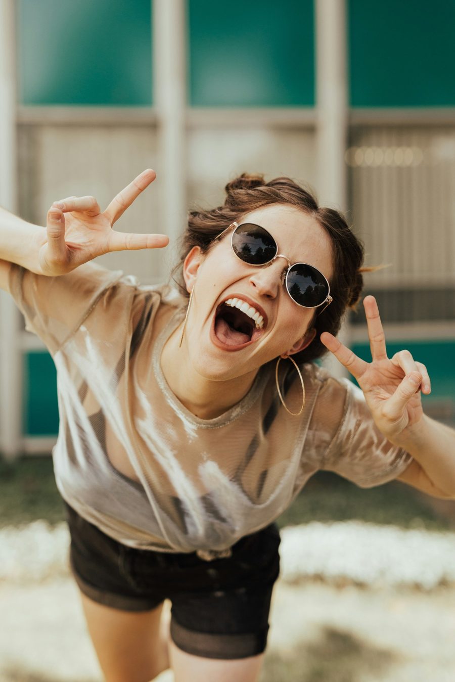 Junge Frau mit Sonnenbrille lacht und zeigt mit beiden Händen das Peace-Zeichen